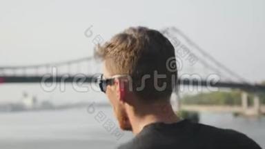 活泼苗条的男人沿着河跑。 年轻的肌肉男在桥附近跑步。 在城市慢跑时，精力充沛的跑步者